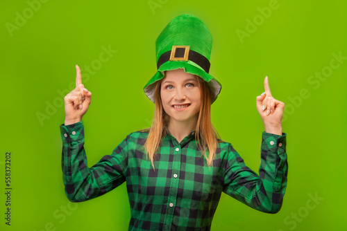 St Patricks Day leprechaun girl pointing index fingers up or holding fingers up, wearing green checkered plaid shirt with green traditional irish hat and looking up isolated on green background.