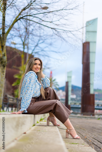 Young woman sitting in a park in the city smiling, lifestyle concept, blue shirt and brown pants