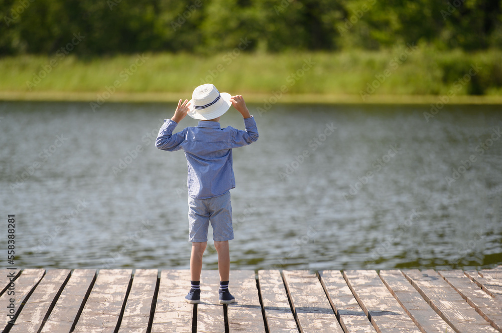A boy in a hat stands on a wooden pier by the lake, looks into the distance and dreams on a summer day