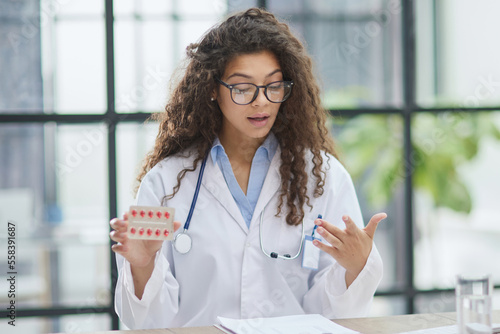 Young pharmacist woman holding pills while sitting at the table in the office