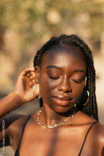 Close-up portrait of a young black female removing her hair from her face whit her eyes closed during sunset