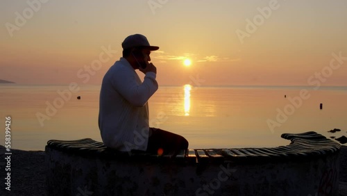 young man wearing hat sitting on beach bench at sunset making long phone call