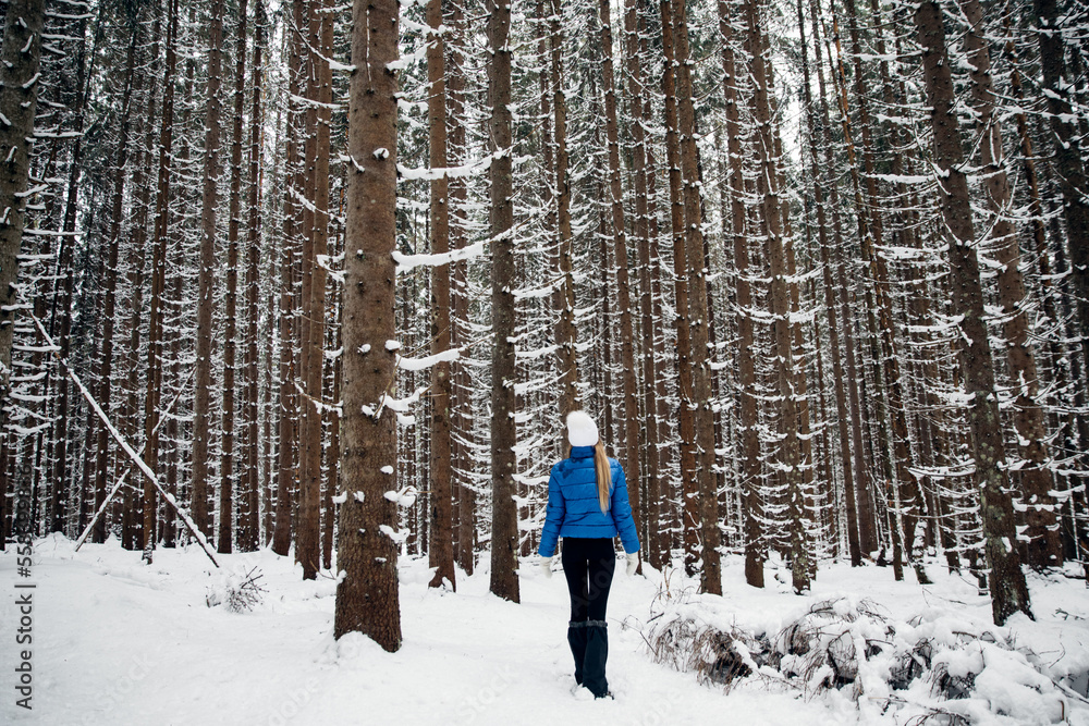 Beautiful woman standing among snowy trees in winter forest and enjoying first snow. Wearing white hat, and blue coat