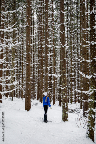 Beautiful woman standing among snowy trees in winter forest and enjoying first snow. Wearing white hat, and blue coat.