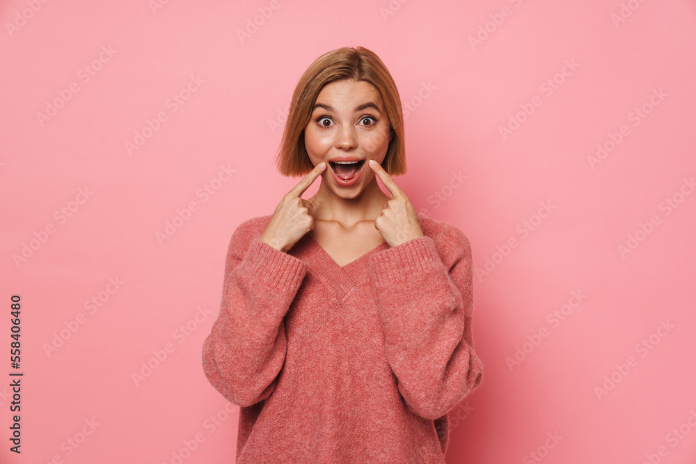 Positive woman screaming and pointing fingers at her cheeks isolated over pink background Positive woman screaming and pointing fingers at her cheeks isolated over pink background