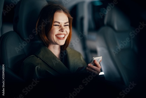a close horizontal portrait of a stylish, luxurious woman in a leather coat sitting in a black car at night in the passenger seat, happily looking at her phone during the trip © SHOTPRIME STUDIO