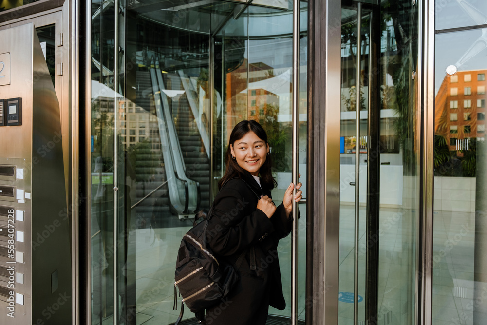 Young asian woman with backpack smiling while standing by doors