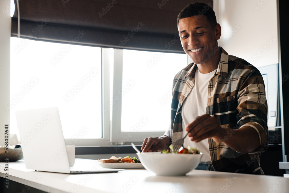 Young positive african man using laptop while having lunch at home Young positive african man using laptop while having lunch at home