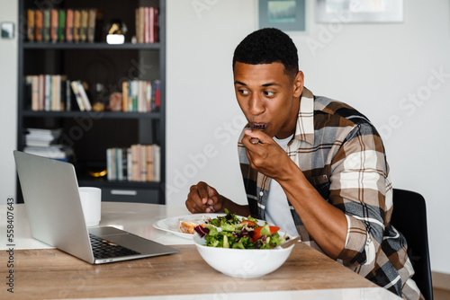 Young african man watching movie on laptop while having lunch at home