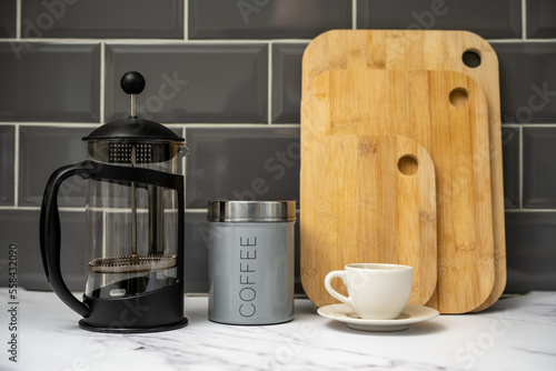 coffee air tight cannister Glass Cafetiere with coffee cup and saucer bamboo chopping boards in the kitchen against a grey tiled background