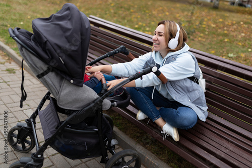 a young woman with a pram in the park listens to music on headphones sitting on a bench