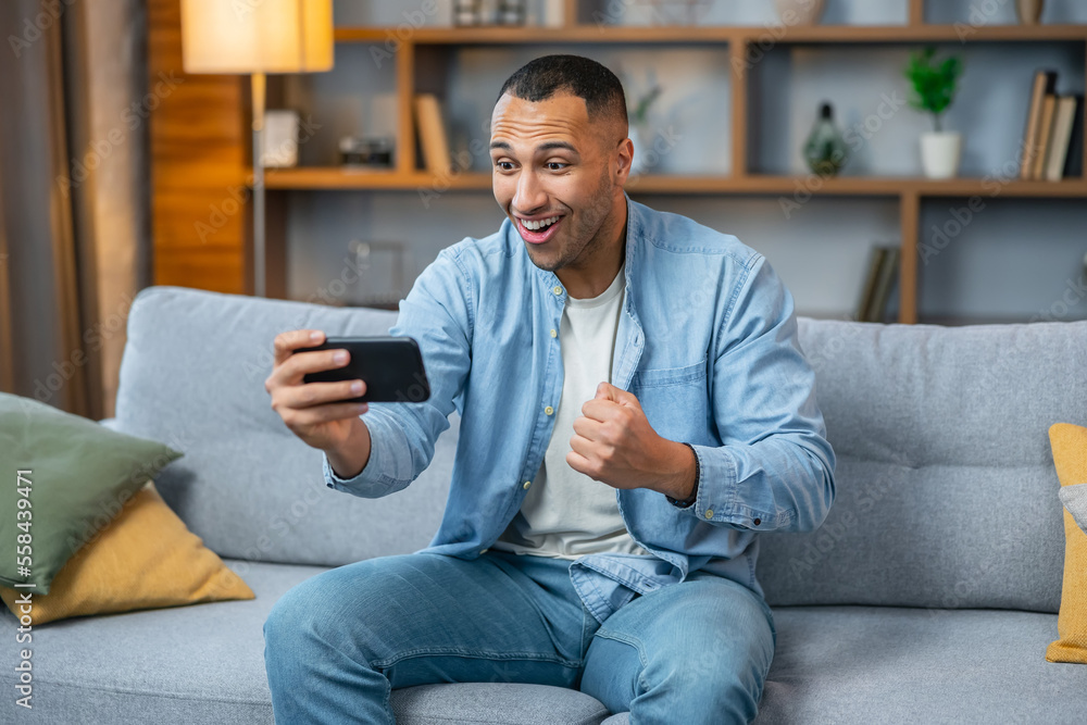 Excited and shocked gamer with headset playing video mobile game online on his smartphone, sitting on sofa, rejoices in victory