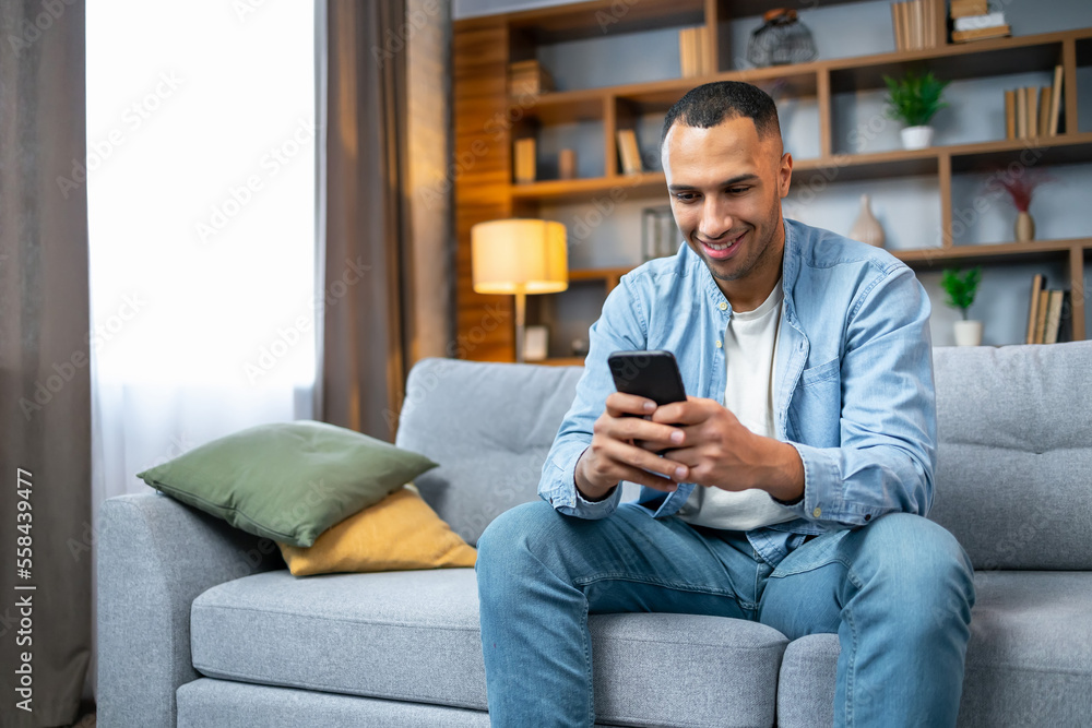 Handsome african american man using smartphone while relaxing sitting on couch at home in living room, browsing internet or messaging with friends, Free Copy Space Handsome african american man using smartphone while relaxing sitting on couch at home in living room, browsing internet or messaging with friends, Free Copy Space