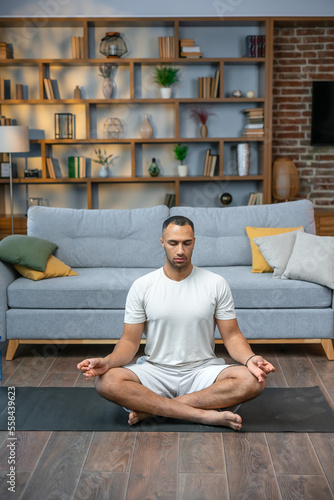 Young man meditating on his living room floor sitting in the lotus position with his eyes closed and an expression of tranquility in a health and fitness concept