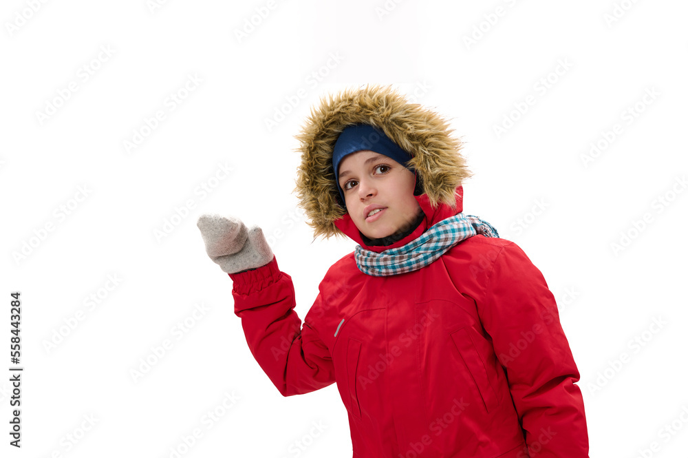 Adorable Middle-Eastern teenage boy, in red down parka with fur hood, blue scarf and woolen mittens, smiles looking at camera, points at copy space for your ads, white background. Cool weather. Winter Adorable Middle-Eastern teenage boy, in red down parka with fur hood, blue scarf and woolen mittens, smiles looking at camera, points at copy space for your ads, white background. Cool weather. Winter