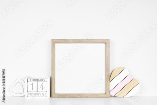 Mock up square wood frame with rustic Valentines Day wooden heart decor and calendar. White shelf against a white wall. Copy space.