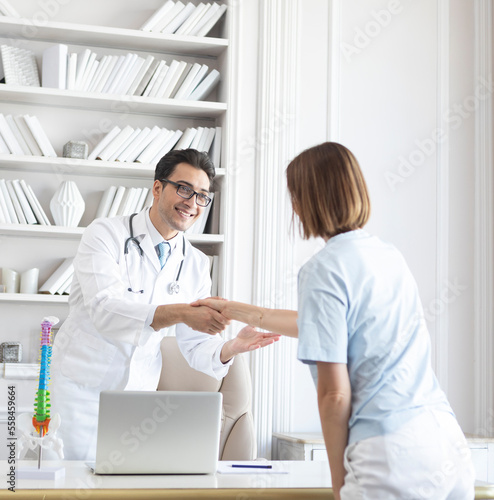Young handsome physician in white medical gown receiving female patient in his modern workplace