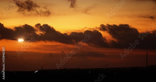 Dramatic orange sky covered with clouds and flying silhouette of airplane that is landing and lights its way with headlights. Beautiful and gorgeous summer sunset, sky with flying airplane silhouette.