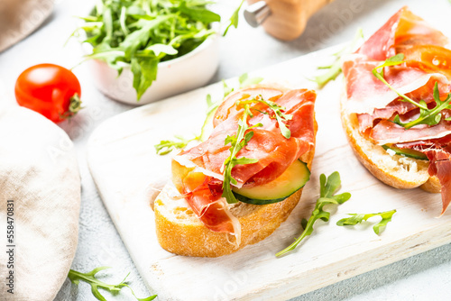 Open sandwiches with cream cheese, prosciutto, cucumber and arugula at white table.