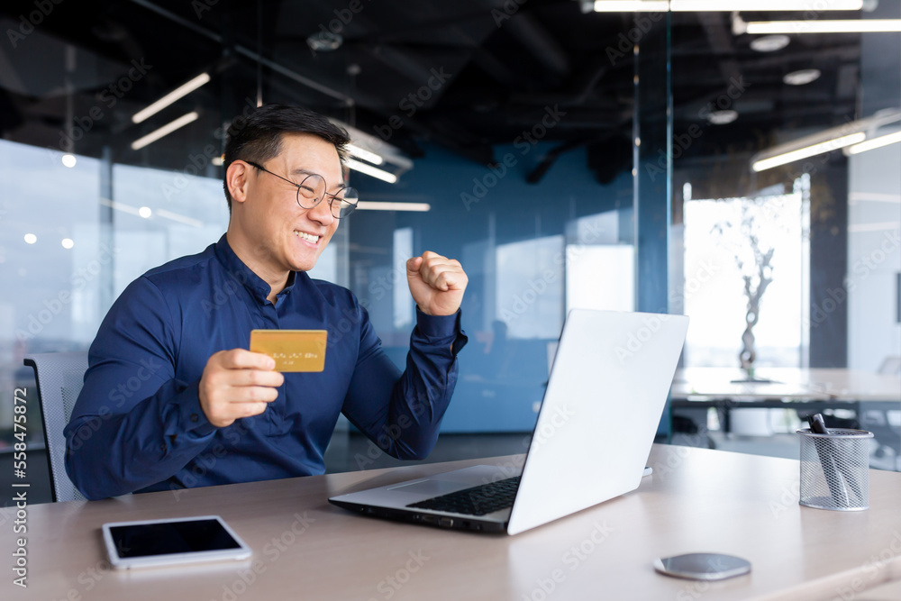 Φωτογραφία Happy and successful businessman in office making online payments inside building, asian man using bank credit card and laptop for online shopping, man working sitting at desk