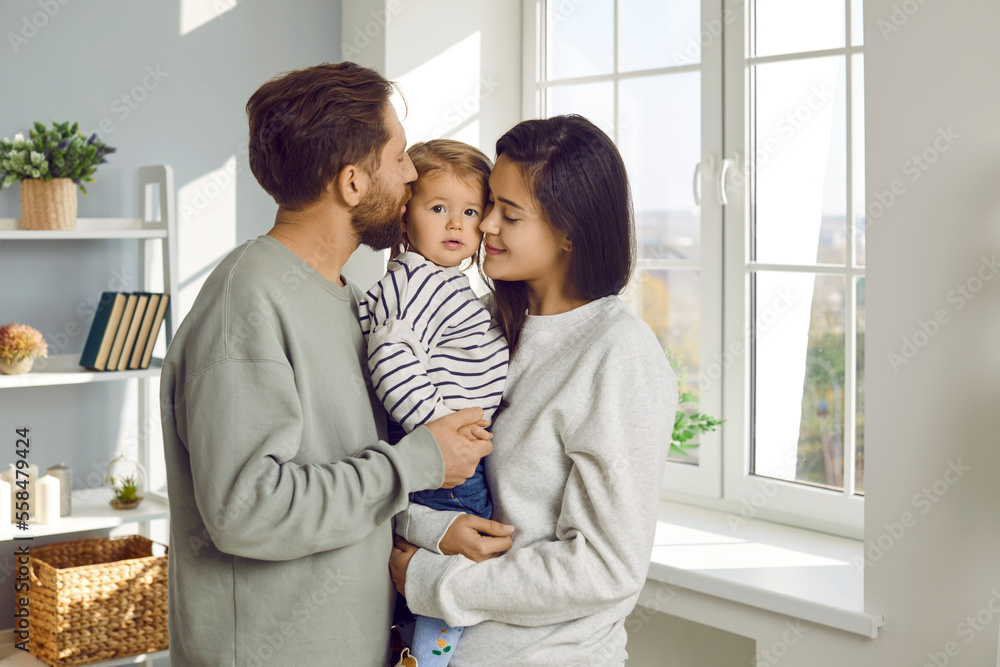 Happy young family. Portrait of young parents kissing their little child, who they are holding together in their arms. Caucasian husband and wife posing with daughter in living room at home. Happy young family. Portrait of young parents kissing their little child, who they are holding together in their arms. Caucasian husband and wife posing with daughter in living room at home.