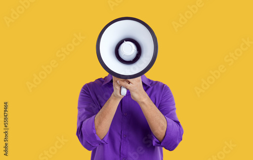Man hiding his face behind loudspeaker makes loud announcement or advertisement on orange background. Unknown man in purple casual shirt holding bullhorn in front of his face. Advertisement concept. photo