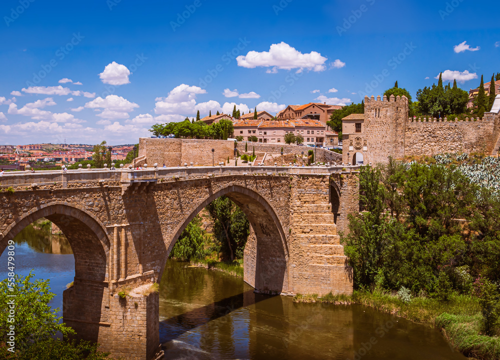 View of medieval bridge in Toledo, Spain, on nice summer day; blue sky with clouds in background; small figures of people on the bridge