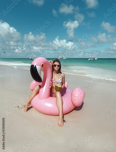 A beautiful sexy amazing young woman on the beach sits on an inflatable pink flamingo and laughs, has a great time, tanned perfect body, long hair, white bikini, fashion accessories, low key photo 