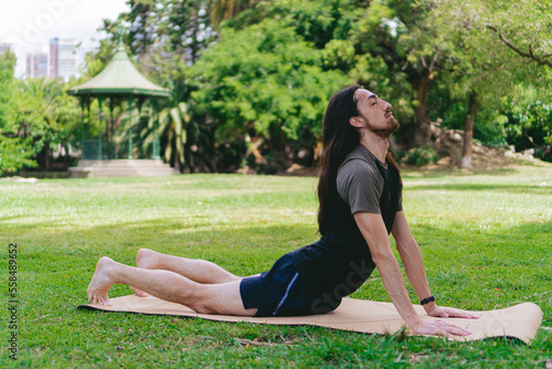 Young, Latino, Hispanic male practicing yoga doing the cobra pose with eyes closed and head up on a mat in a green field.