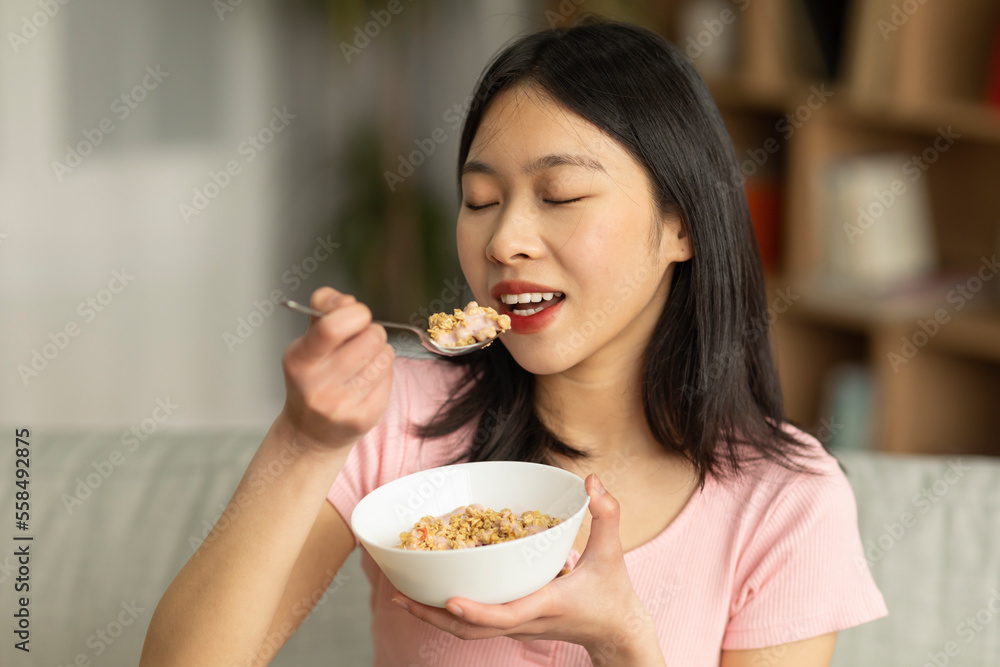 Portrait of young asian lady having cereal with milk while sitting on sofa at home, enjoying healthy meal Portrait of young asian lady having cereal with milk while sitting on sofa at home, enjoying healthy meal