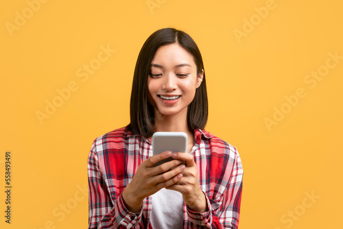 Young asian woman using smartphone for messaging  smiling lady texting on cellphone  yellow background  free space