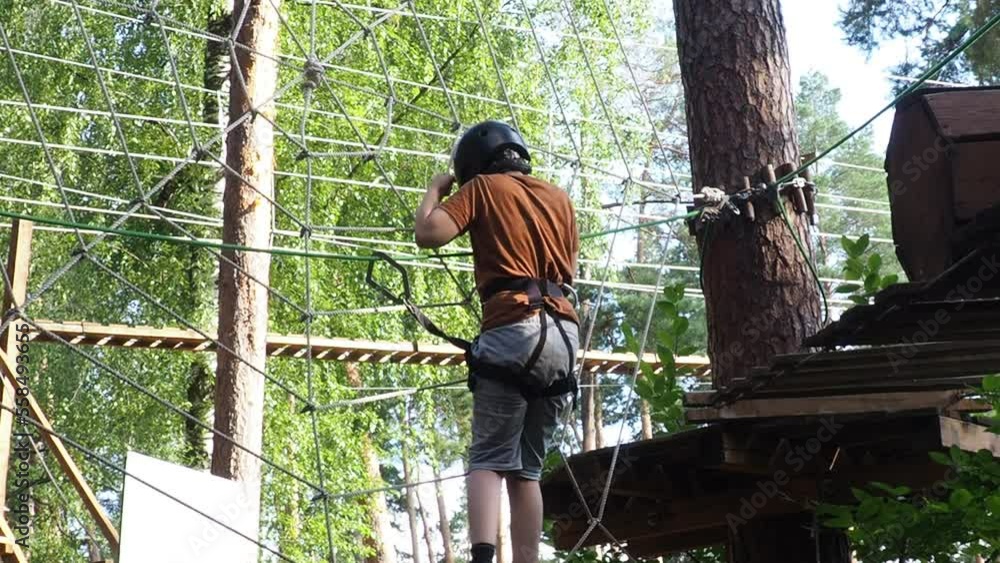 Portrait of cute little boy walk on a rope bridge in an adventure rope park. Child in forest adventure park. Kids climb on high rope trail. Agility and climbing outdoor amusement center for children. Portrait of cute little boy walk on a rope bridge in an adventure rope park. Child in forest adventure park. Kids climb on high rope trail. Agility and climbing outdoor amusement center for children.