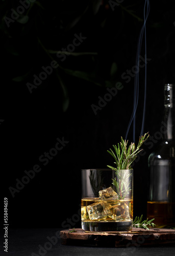 Whiskey, scotch or bourbon glass with smoking rosemary, ice cubes on black background