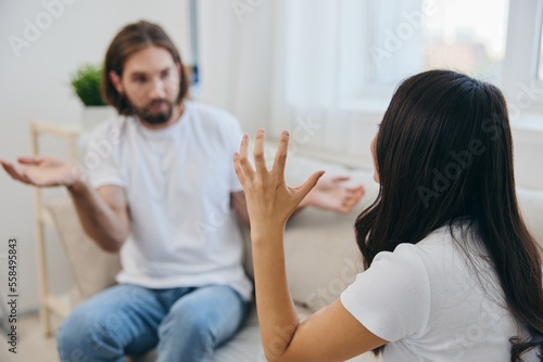 A man and a woman sit on the couch at home in white T-shirts and chatting scandalously do not understand each other. A quarrel in the family of two spouses and aggression