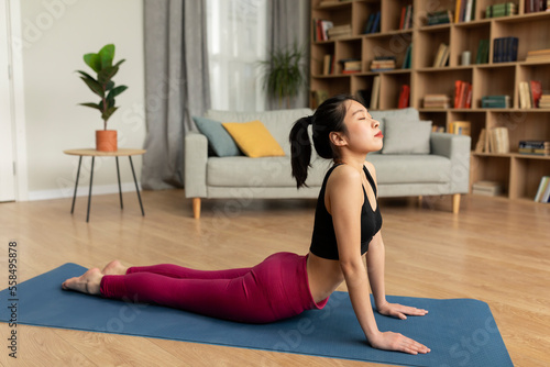 Fit korean lady doing yoga cobra pose or pilates, working out on mat in living room interior, copy space