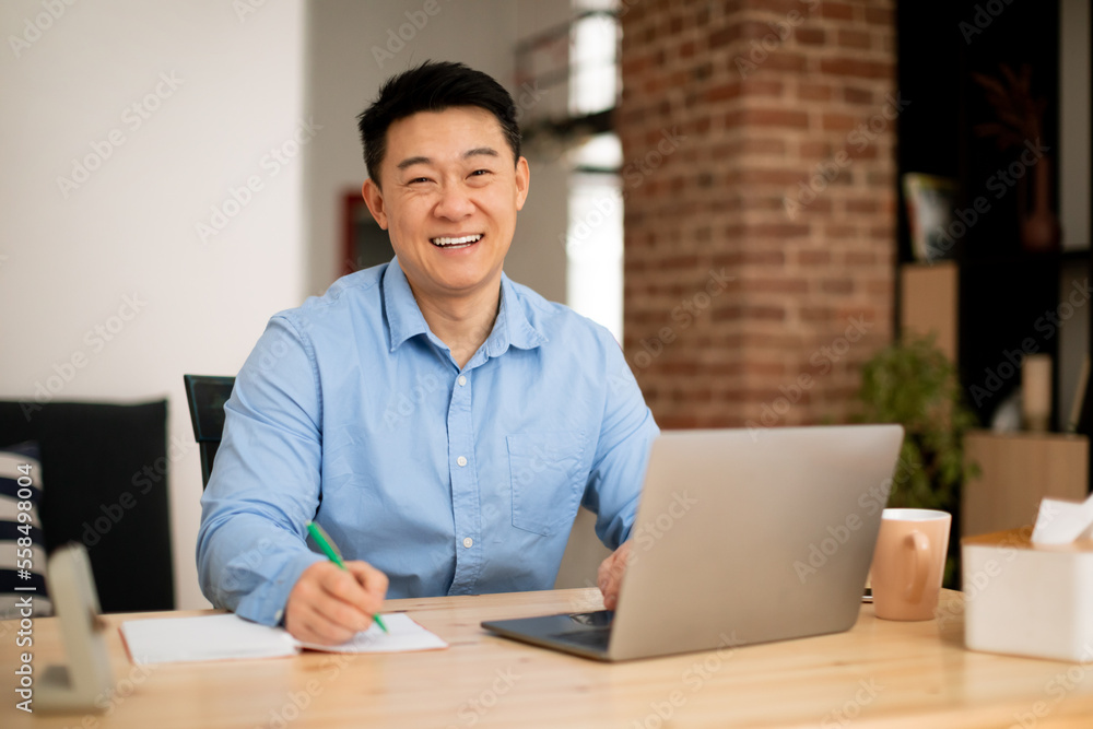Happy asian middle aged man working on laptop and taking notes in notebook, smiling at camera, free space Happy asian middle aged man working on laptop and taking notes in notebook, smiling at camera, free space