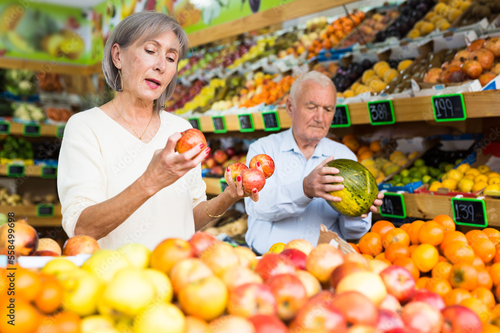 Caucasian mature woman and man selecting ripe fruits in greengrocer.