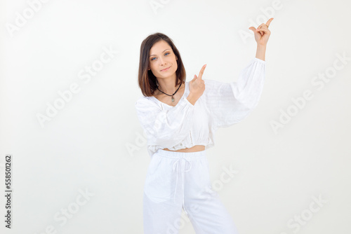 Portrait of young woman standing, raising hands, pointing with index fingers, looking at camera on white background.