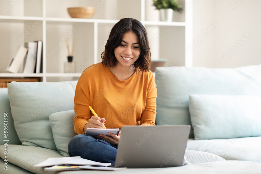 Smiling Young Arab Female Student Using Laptop At Home And Taking Notes Smiling Young Arab Female Student Using Laptop At Home And Taking Notes