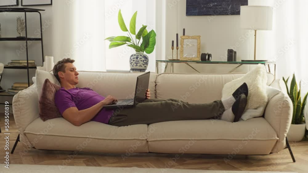 A man works with a computer at home while lying on the sofa. Comfortable remote work online. A freelancer performs tasks. Handsome Caucasian young man working on laptop. High quality 4k footage A man works with a computer at home while lying on the sofa. Comfortable remote work online. A freelancer performs tasks. Handsome Caucasian young man working on laptop. High quality 4k footage