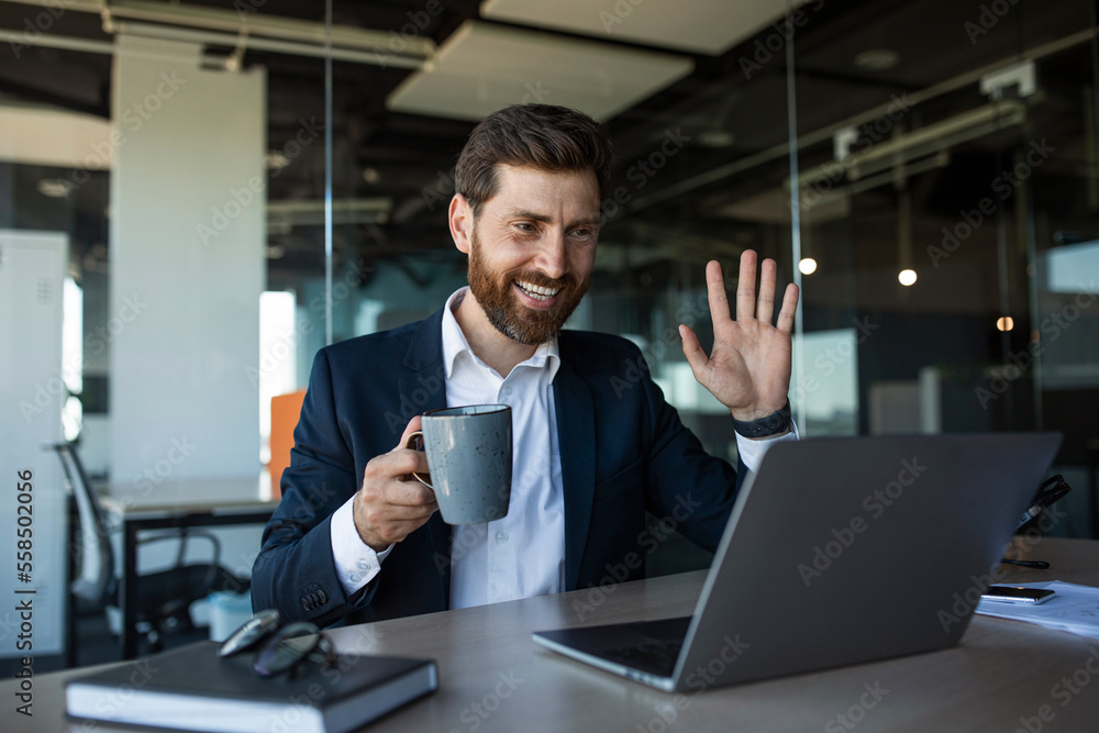 Smiling middle aged businessman waving hand at laptop webcamera, having meeting in office interior, free space