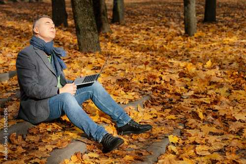 Side view of middle-aged man sitting with closed eyes on stairs covered with yellow leaves in autumn, holding laptop.