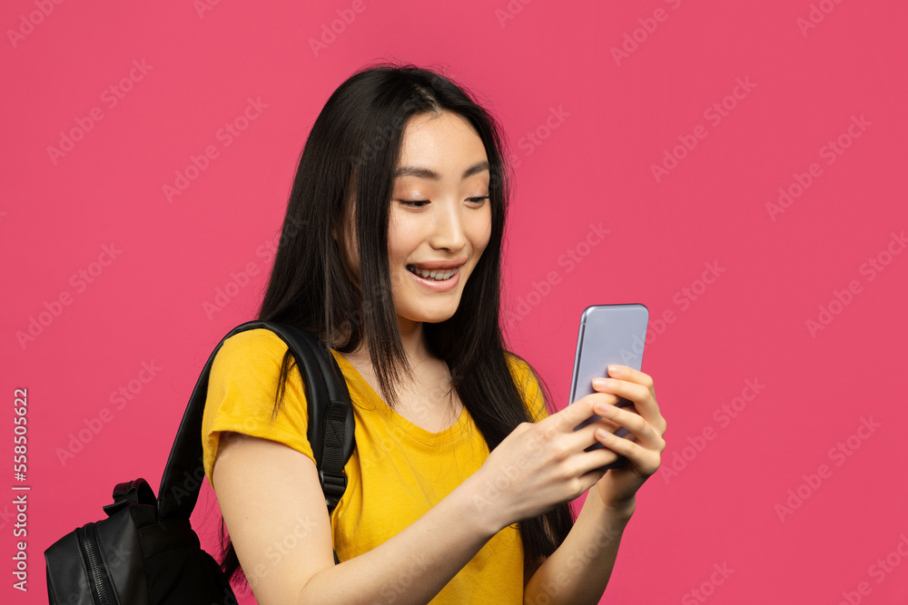 Happy asian female student using smartphone, texting online while standing with backpack on pink background, copy space Happy asian female student using smartphone, texting online while standing with backpack on pink background, copy space