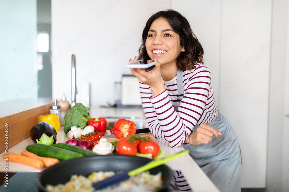 Positive young arab woman recording voice message while cooking