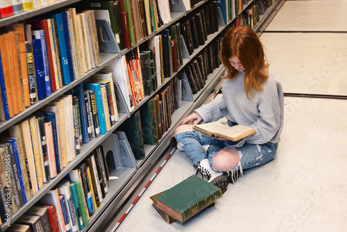 Teen Redhead Girl Sitting With Pile of Books in Library photo