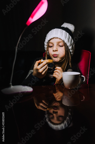 Blackout in Ukraine. Ukrainian beautiful girl, a child in warm clothes, a hat, a blanket, a blanket, sits at a table in a cold room in the dark with a lamp, eats a sandwich, drinks hot tea from a cup.