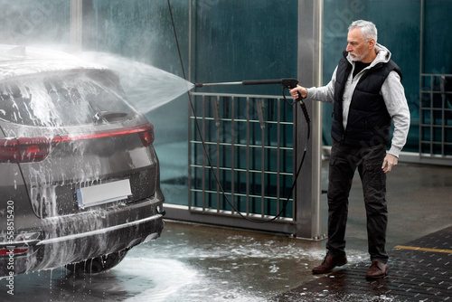 Full length Senior man washes off the foam from his car with water from pressurized hose at car wash photo