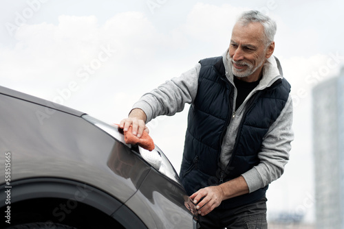 Smiling Caucasian active senior man wiping headlights of his luxury clean in outdoors parking lot