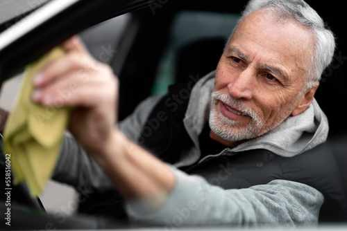 Close-up portrait of handsome positive white-bearded man driver, wiping the car's side mirror