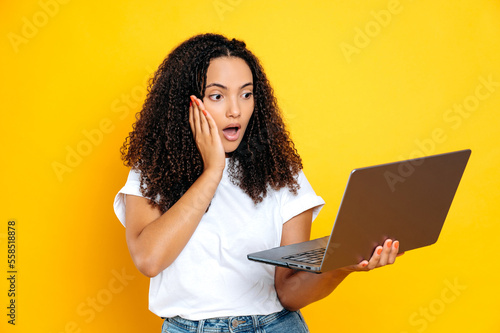 Excited shocked brazilian or hispanic young curly woman, wearing basic white t-shirt, holding open laptop looking in amazement at it, standing on isolated orange background, surprised expression